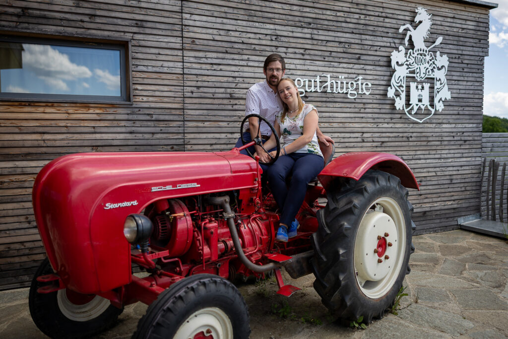 Paar auf einem roten Oldtimer-Traktor vor der Holzfassade mit Gut-Hügle-Schriftzug – Engagement-Shooting im Gut Hügle, Hochzeitsfotograf Ravensburg